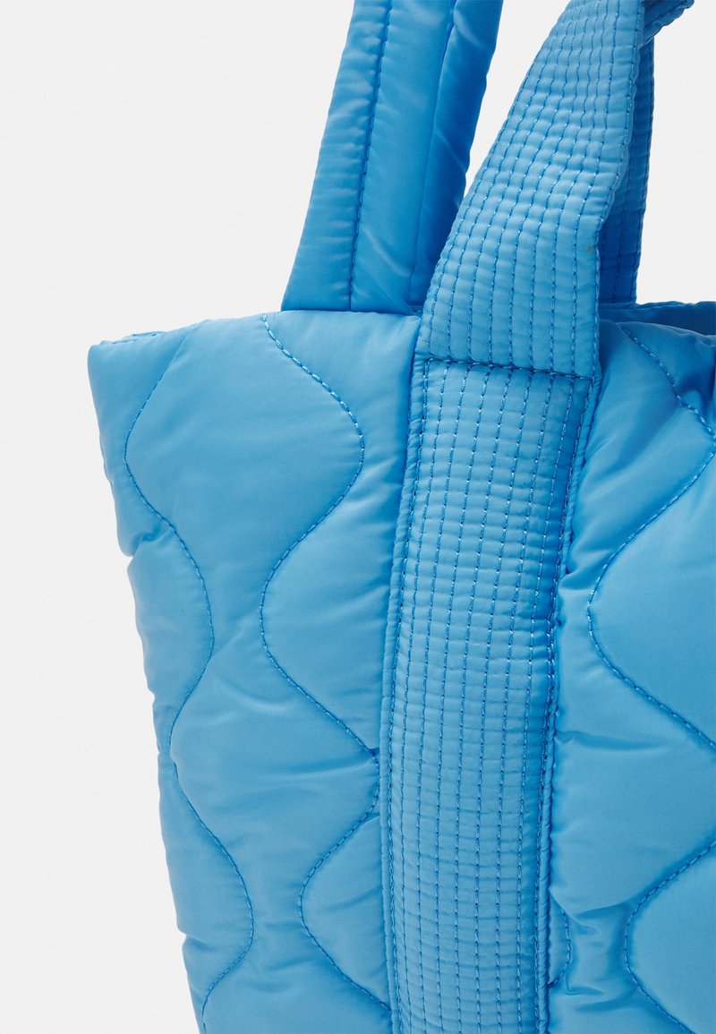 Niebieska torba typu tote, pikowana, z miękką teksturą, z ukośnymi wzorami stębnowania oraz szerokimi, płaskimi uchwytami. Krawędzie są zaokrąglone.