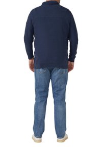 Marineblauer Strickpullover mit geripptem Kragen und Schulterdetail, kombiniert mit hellblauen Denim-Jeans und weißen Turnschuhen. Legerer Schnitt.