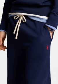 Navy sweatpants with a drawstring waist, featuring a small red logo on the pocket and a smooth texture. Accented with a light blue layer underneath.