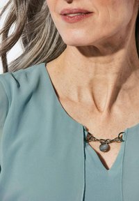 Helle blaue Bluse mit V-Ausschnitt und metalischer Kette mit einem runden Charm, vor glatter Haut und grauem Haar.
