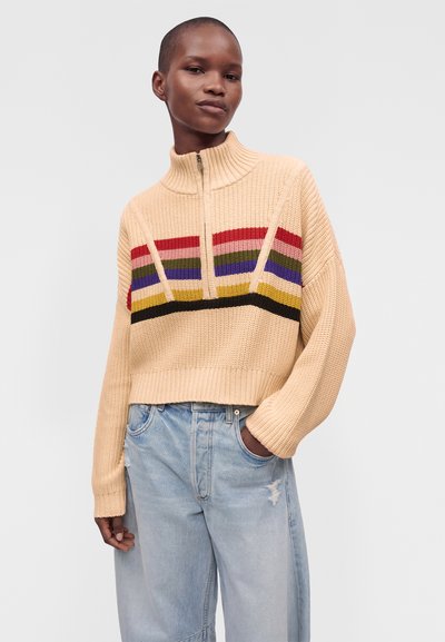 Personne portant un pull en maille beige avec des rayures horizontales colorées et un col à demi-fermeture éclair, associé à un jean déchiré bleu clair.