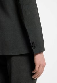 Primer plano de una persona vistiendo un traje gris oscuro con tres botones negros en la manga y su mano derecha relajada a un lado.