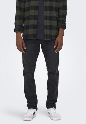 Homme portant un jean noir, des baskets blanches, une chemise noire et une chemise à carreaux vert et noir, debout devant un fond uni.