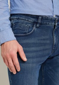 Man som bär mid-blå jeans och en ljusblå texturerad långärmad skjorta, hand vilande vid sidan nära fickan.