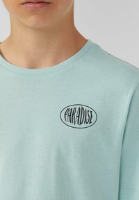 Lichtblauwe katoenen t-shirt met een ronde halslijn met een zwart ovale "PARADISE" logo. Zachte textuur, aansluitend ontwerp en korte mouwen.