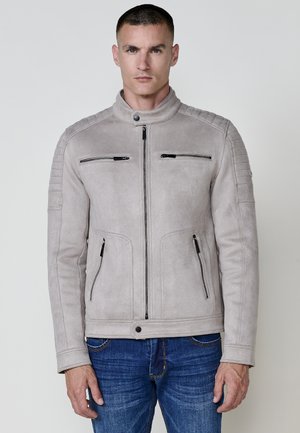 Chaqueta de ante gris con un cuello alto, cierre frontal con cremallera, dos cremalleras en el pecho, bolsillos laterales y acentos texturizados en los hombros; combinada con jeans azules.