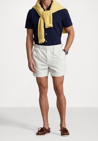 Navy short-sleeve shirt, cream shorts, and yellow cable-knit sweater draped over shoulders; brown boat shoes complete the look.