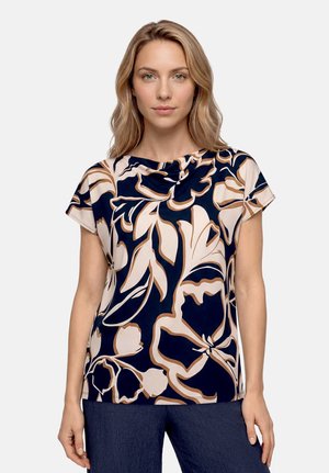 Femme aux cheveux blonds portant un haut bleu marine avec de grands motifs floraux beiges et blancs et un pantalon bleu foncé, debout devant un fond uni.