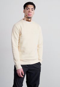 Pulover crem deschis din bumbac, cu logo embossat, guler rotund și manșete ribbate. Purtați cu pantaloni cargo negri pe un fundal neutru.