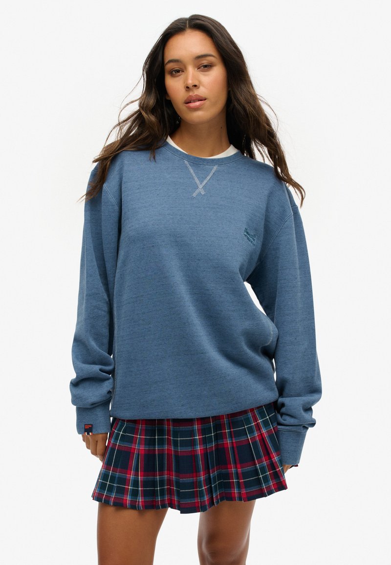 Sudadera azul de cuello redondo con un acabado texturizado, que presenta un detalle de cruz cosido en el cuello. Combinada con una falda de cuadros plisada en tonos azul marino y rojo.