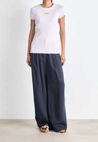 Ljusrosa åtsittande t-shirt med orange logotyp, i kombination med lös mörkgrå byxor och bruna sandaler med vita remmar.