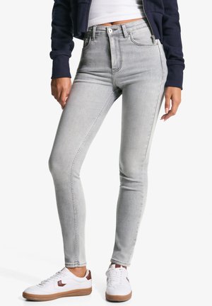 Femme portant un jean skinny gris clair, une veste bleu marine, une chemise blanche et des baskets blanches avec des détails marron, debout, les mains détendues le long du corps.