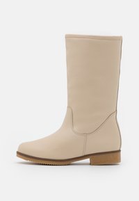 Bota de cuero beige hasta la rodilla con puntera redondeada, textura suave, detalle de costura lateral y un tacón bajo y cuadrado. Suela de goma para un mejor agarre.