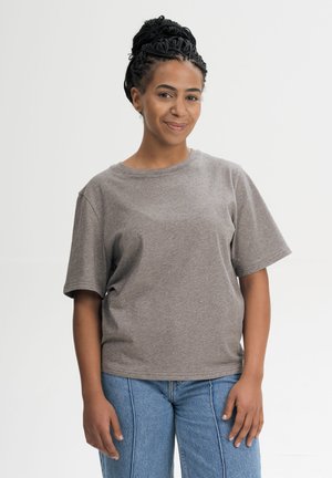 Vrouw met gevlochten haar die een grijs T-shirt met korte mouwen en een blauwe spijkerbroek draagt, staat en glimlacht tegen een effen achtergrond.