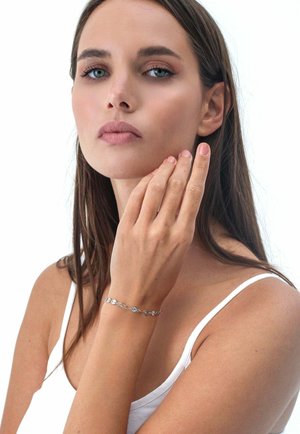 Silver bracelet with circular decorations displayed on a hand. Model with long brown hair wearing a white tank top, clear skin, and minimal makeup.