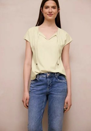 Ung kvinde iført en lysegul bluse med korte ærmer og snøre samt blå højtaljede jeans, stående mod en ensfarvet baggrund.