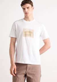 Hvit bomulls t-skjorte med et grafisk motiv av stiliserte lepper i beige toner, med en prikkete tekstur bak bildet og merkevare over.