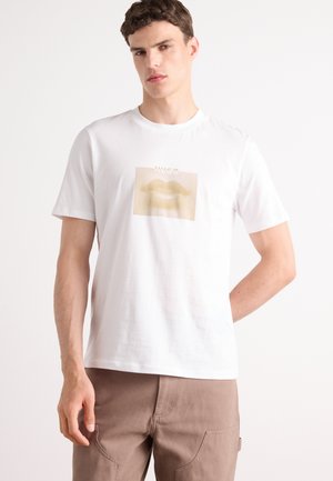 Hvit bomulls t-skjorte med et grafisk motiv av stiliserte lepper i beige toner, med en prikkete tekstur bak bildet og merkevare over.