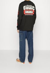 Camisa negra de manga larga con estampados gráficos en la parte posterior, combinada con jeans de denim azul sueltos y zapatillas con acentos naranjas.