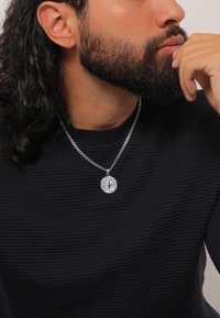 Sidabrinis kompaso pakabukas ant grandinėlės, nešiojamas ant tekstūruoto juodo riboto megztinio. Detalėje pavaizduotas įgasdytas kompaso raštas.