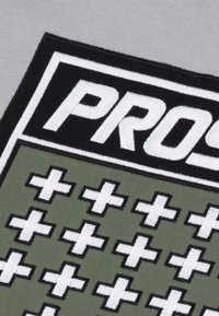 Naszywka z haftowanym słowem "PROS" w białym kolorze na czarnym tle, z zielonym tłem z wzorem białych krzyżyków obramowanych na czarno.