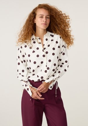 Jonge vrouw met lang krullend rood haar, gekleed in een witte blouse met zwarte polkadots en bordeauxrode hoge taille broek.