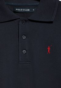 Zwarte polo met drie knopen. Kenmerkt zich door een gestructureerde stof en een klein rood logo van een golfer op de linkerkant van de borst. Etiket leest "POLO CLUB."