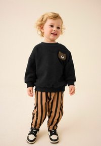 Sweat-shirt noir avec un détail de poche en forme d'ours, associé à un pantalon rayé beige et marron. Des baskets blanches et noires complètent la tenue.