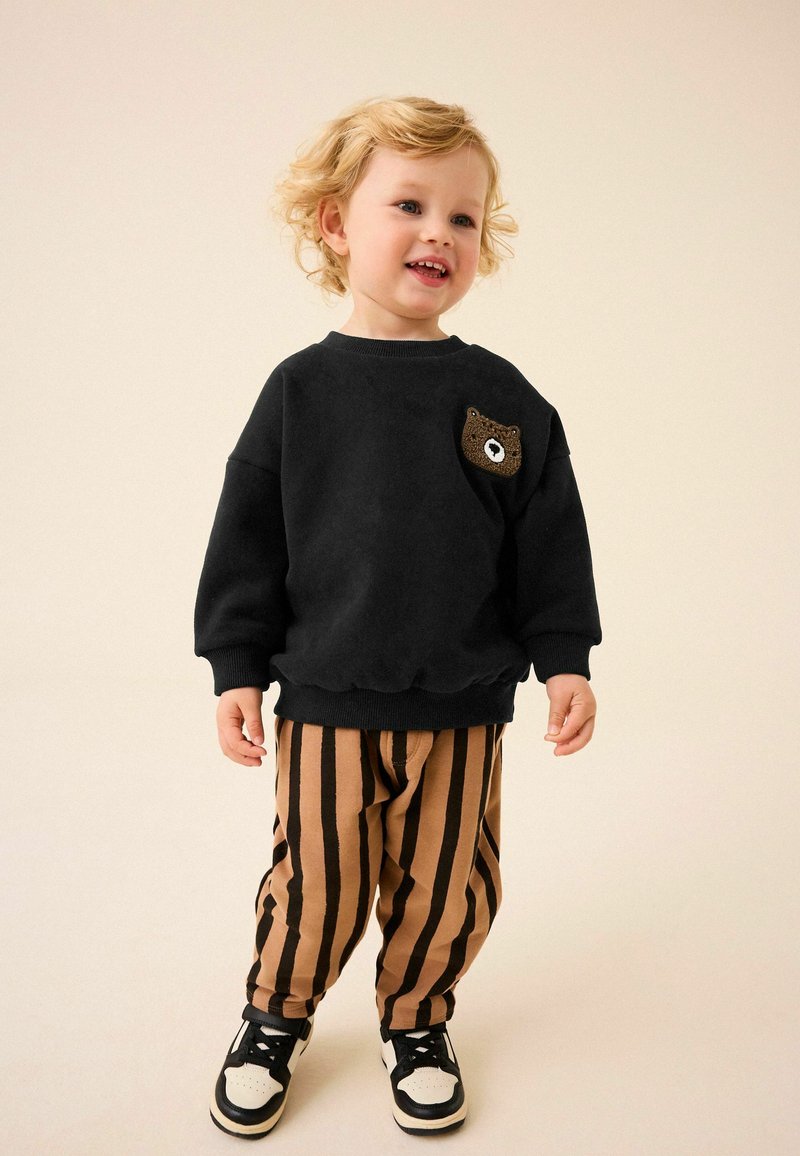Sweat-shirt noir avec un détail de poche en forme d'ours, associé à un pantalon rayé beige et marron. Des baskets blanches et noires complètent la tenue.