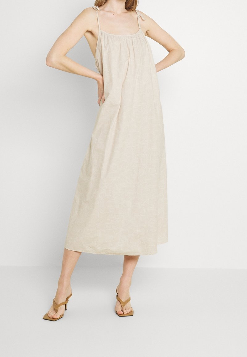 Robe maxi sans manches beige avec un buste froncé, de fines bretelles et une texture subtile. Associée à des sandales à talons marron.