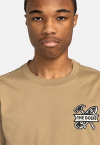Beige bomulds t-shirt med et sort grafisk print af en hammer og to fisk, samt teksten "THE DOERS" på venstre bryst.