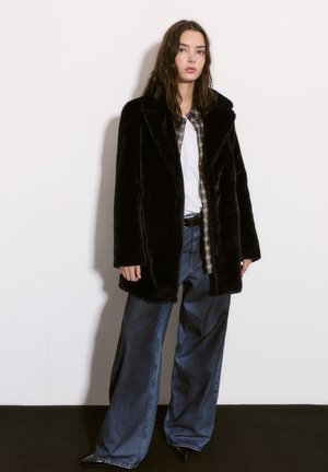 Cappotto in finta pelliccia nera con ampi revers, indossato sopra una camicia a quadri e una maglietta bianca, abbinato a jeans a gamba larga blu chiaro e scarpe nere a punta.