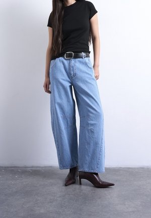 Mujer con camiseta negra, vaqueros de pierna ancha azul claro con cinturón negro y botas de tacón alto puntiagudas de color marrón oscuro, de pie sobre suelo gris.