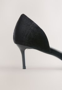 Escarpin à talon aiguille en velours noir à bout pointu avec un talon fin et élégant et un détail ouvert sur le côté, sur fond neutre.
