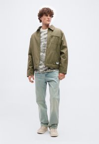 Νέος άνδρας με σγουρά μαλλιά, φορώντας πράσινο καμελί μπουφάν, γκρι παραλλαγής πουκάμισο, ανοιχτό μπλε τζιν και μπεζ αθλητικά παπούτσια, στέκεται μπροστά σε απλό φόντο.