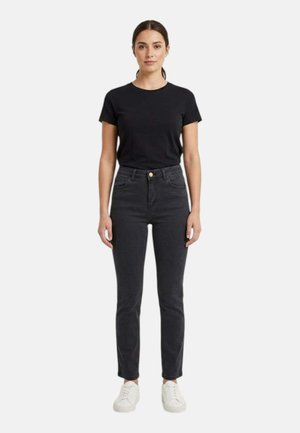 Donna in piedi che indossa una t-shirt nera a maniche corte, jeans neri a vita alta e sneakers bianche su sfondo semplice.