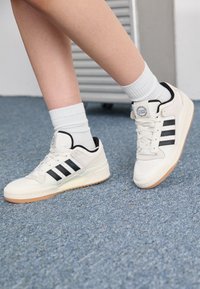 White sneakers with three black stripes, textured rubber sole, round toe, and padded collar, worn with ribbed white socks.
