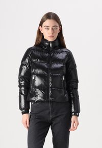 Zwarte pufferjack met een hoge kraag, glanzende afwerking, horizontaal geruit ontwerp en ritszakken aan de zijkant. Gecombineerd met donkere jeans.