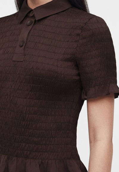 Gros plan d'une personne portant un chemisier texturé marron foncé à manches courtes avec un col et trois boutons sur la patte de boutonnage avant.