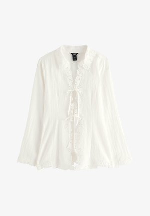 Blusa bianca a maniche lunghe in tessuto leggero, con bordatura in pizzo floreale, scollo con fiocco e orlo smerlato. Dettagliata con accenti ricamati.