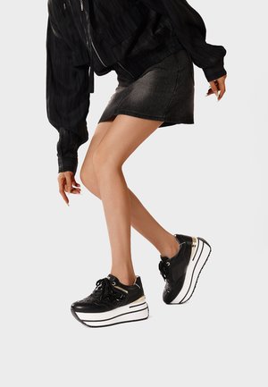 Donna che indossa sneakers nere con suola platform, una gonna corta scura e una giacca nera, posando davanti a uno sfondo chiaro e uniforme.