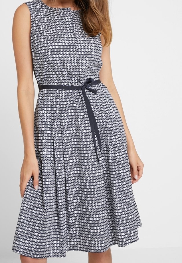 Femme portant une robe sans manches à motifs noir et blanc, longueur genou, avec une fine ceinture noire nouée à la taille, sur fond blanc.