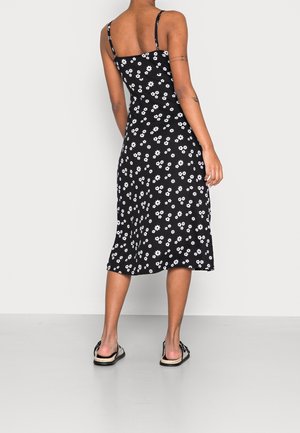 Robe midi noire à motifs floraux blancs de marguerites, bretelles spaghetti, tissu lisse et ourlet évasé, portée avec des sandales plates.