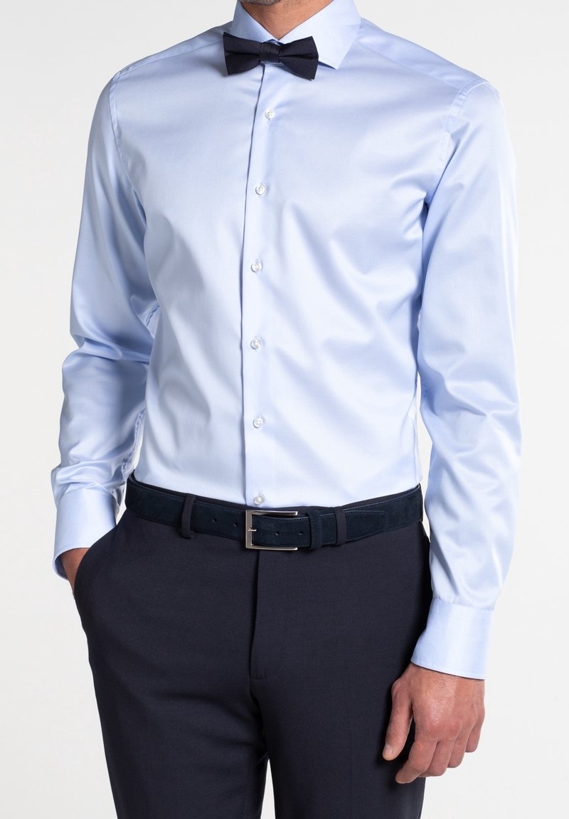 Homme portant une chemise bleu clair avec un nœud papillon noir, un pantalon noir et une ceinture noire, debout avec une main dans la poche.