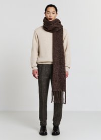 Brauner Wollschal, kunstvoll um den Hals drapiert, mit strukturierter Strickoptik und fransigen Enden, kombiniert mit einem cremefarbenen Pullover und gemusterten Hosen.