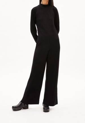 Kvinde iført en sort jumpsuit med lange ærmer og vide ben samt sorte studdede platform klipklapper, stående med hænderne bag sin ryg.