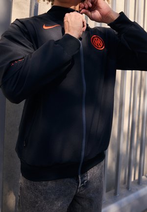 Person, die eine schwarze Jacke mit orangefarbenem Nike-Logo und Emblem zumacht, steht neben vertikalen Metallstäben, durch die Sonnenlicht Schatten wirft.