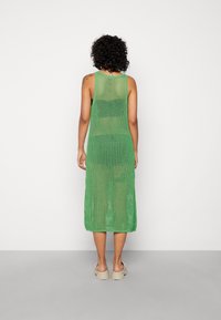 Abito lavorato a maglia verde con un taglio di lunghezza media, caratterizzato da un design trasparente e a giorno e spalline tipo canotta. Indossato con sandali chunky di colore chiaro.