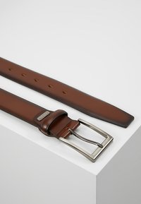 Brun lærrem med en rektangulær sølvfargede metallspenne, med hull langs lengden for størrelsesjustering. Glatt tekstur og solid design.