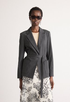Vrouw gekleed in een donkergrijze gebonden blazer, beige top, plissérok met zwart schetspatroon, donkere zonnebril en zilveren hoepeloorbellen.
